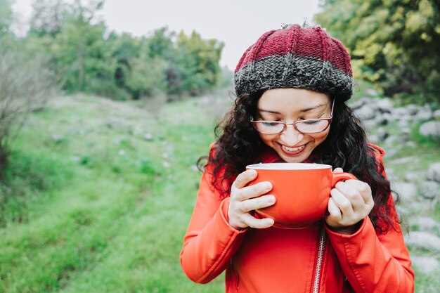 Donna bruna riccia, che indossa una giacca di pelle rossa e un berretto di lana, bevendo caffè caldo in natura.