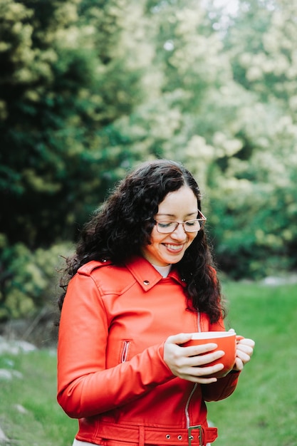 Donna bruna riccia sorridente, che indossa una giacca di pelle rossa e tiene una tazza rossa con caffè caldo