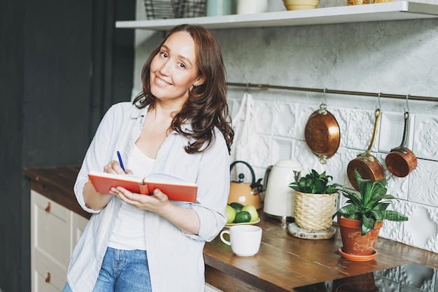 Donna bruna sorridente adulta in appunti casuali nel libro quotidiano con una tazza di tè in cucina a casa