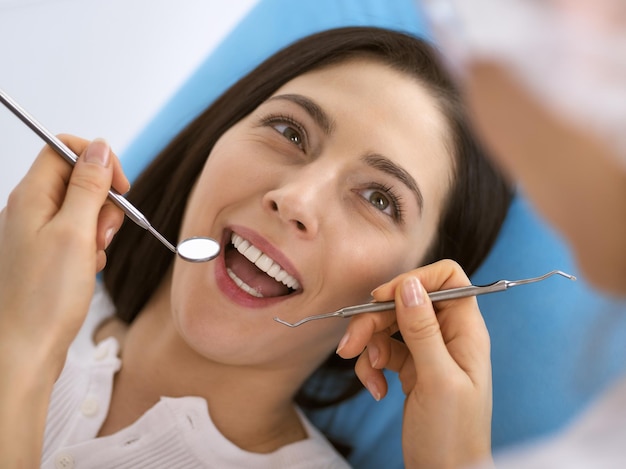 Donna bruna sorridente che viene esaminata dal dentista presso la clinica dentale. Mani di un medico che tiene strumenti dentali vicino alla bocca del paziente. Denti sani e concetto di medicina.