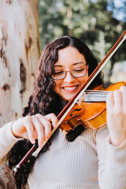 Donna bruna sorridente con gli occhiali che suonano il violino fuori nel bosco Verticale