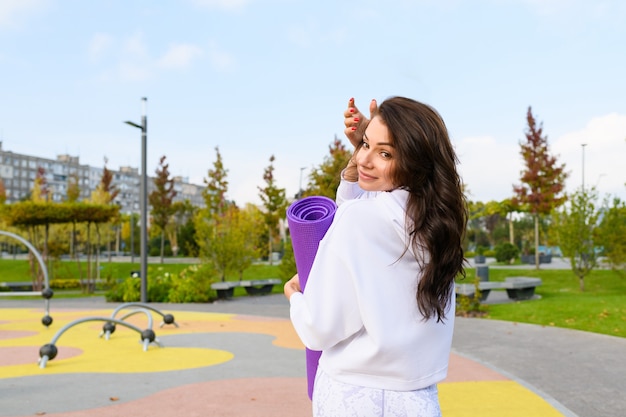 Donna bruna sportiva in felpa con cappuccio bianca tenere tappetino viola al parco giochi del parco cittadino, allenamento, fitness, stretching all'aperto