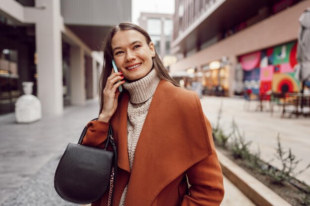 Donna brunetta carina in cappotto marrone che parla al telefono in città