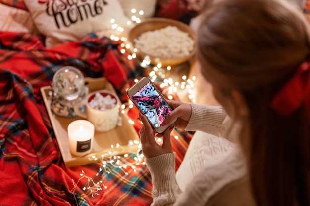 Donna candida senza volto che tiene in mano lo smartphone e riprende video di cibi e bevande e decorazioni per il blog. divertimento rilassante in campeggio in un letto accogliente durante la notte di buon Natale e buon capodanno