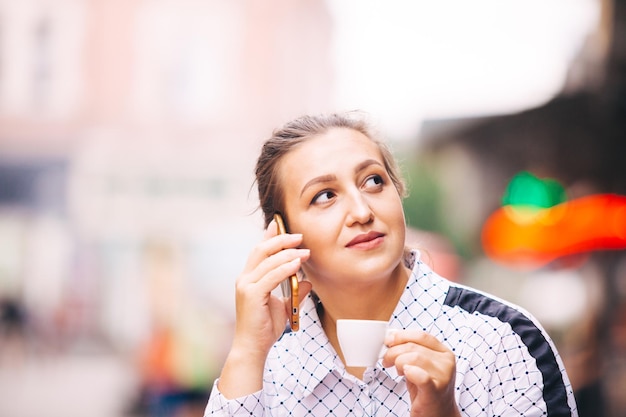 Donna carina in camicetta bianca tiene una tazza di caffè parlando al telefono e distoglie lo sguardo in città