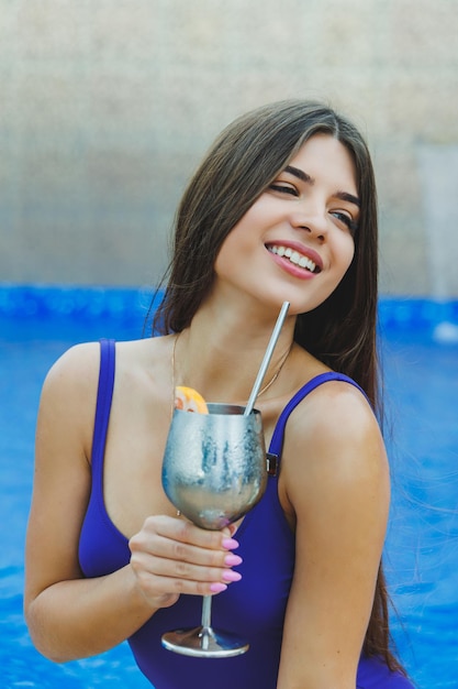 Donna carina in costume da bagno seduta a bordo piscina e bere un delizioso cocktail e prendere il sole a bordo piscina durante le vacanze estive