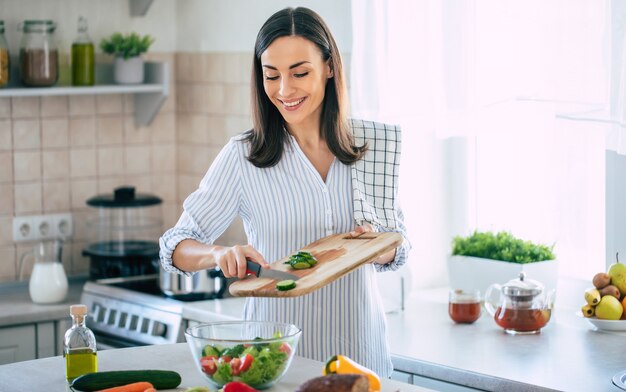 Donna carina sorridente felice sta preparando un'insalata vegana fresca e sana con molte verdure in cucina a casa e sta provando una nuova ricetta