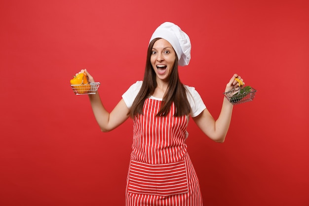 Donna casalinga chef cuoco panettiere in grembiule a righe bianco t-shirt toque cappello chef isolato su sfondo rosso parete. La donna tiene il cetriolo verde del peperone giallo nel carrello di spinta della drogheria Mock up copy space concept