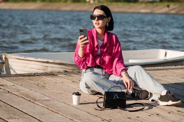 Donna castana alla moda che prende selfie con il cellulare che si siede sul molo di legno sopra lo sfondo del lago in una giornata di sole