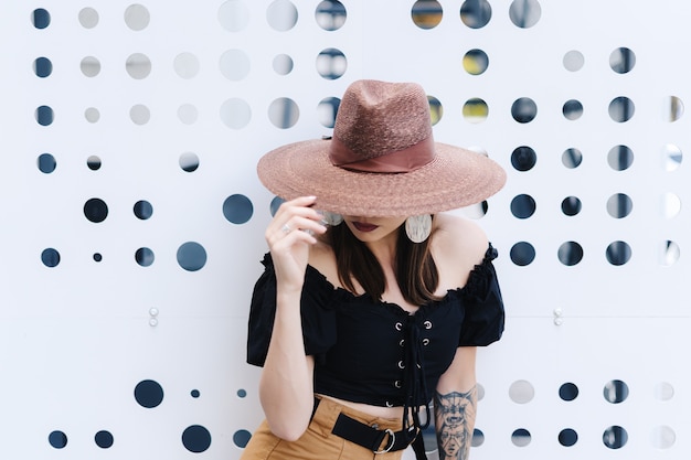 Donna castana alla moda con capelli lunghi, cappello di vimini grande alla moda che indossa, posa contro un muro bianco con buchi.