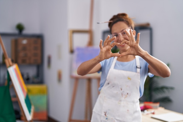 Donna castana che dipinge presso lo studio d'arte sorridendo innamorato mostrando il simbolo del cuore e la forma con le mani. concetto romantico.