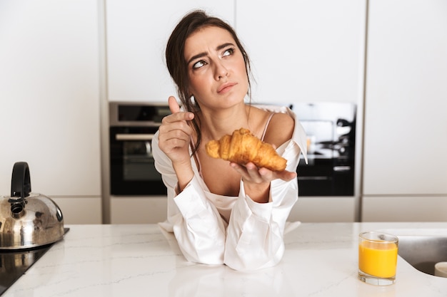 donna castana che indossa abiti di seta che mangia croissant, durante la colazione in cucina a casa