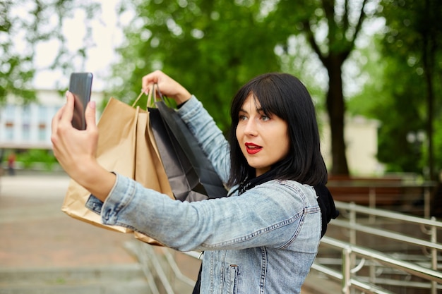 Donna castana che tiene i sacchetti della spesa di carta e si fa selfie con lo smartphone nel parco, ragazza dopo lo shopping.