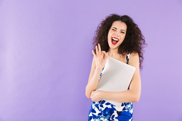 Donna castana con capelli ricci in vestito che sorride e che tiene computer portatile in mani