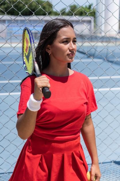 Donna castana con racchetta e palla in piedi vicino al recinto del campo da tennis durante la pausa durante l'allenamento