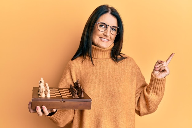 Donna castana di mezza età che tiene la scacchiera sorridendo felice indicando con la mano e il dito di lato