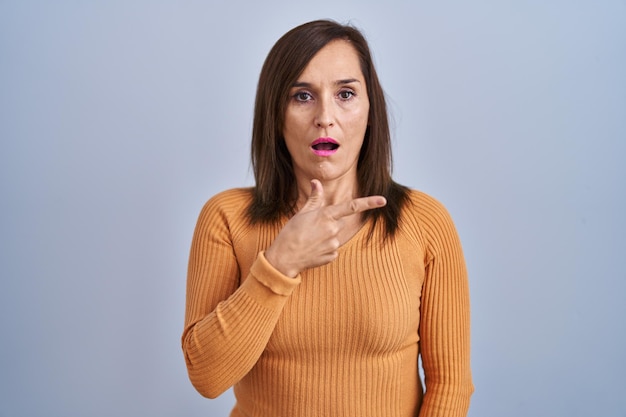 Donna castana di mezza età in piedi che indossa un maglione arancione sorpreso puntando con il dito verso l'espressione stupita della bocca aperta laterale