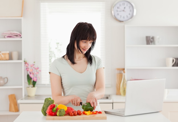 Donna castana felice che cucina mentre rilassandosi con il suo computer portatile