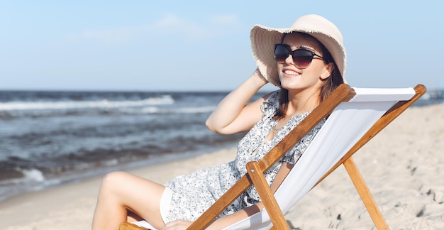 Donna castana felice che indossa occhiali da sole e cappello che si rilassa su una sedia a sdraio in legno sulla spiaggia dell'oceano.