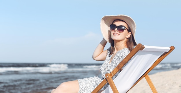 Donna castana felice che indossa occhiali da sole e cappello che si rilassa su una sedia a sdraio in legno sulla spiaggia dell'oceano.