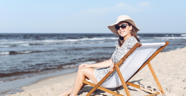 Donna castana felice che indossa occhiali da sole e cappello che si rilassa su una sedia a sdraio in legno sulla spiaggia dell'oceano