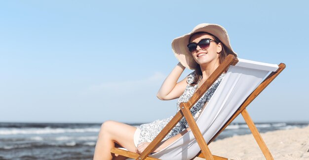 Donna castana felice che indossa occhiali da sole e cappello che si rilassa su una sedia a sdraio in legno sulla spiaggia dell'oceano