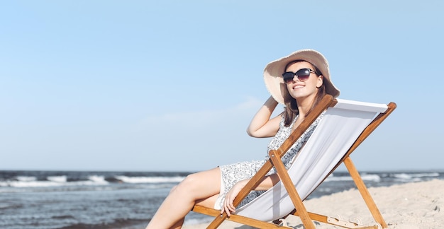 Donna castana felice che indossa occhiali da sole e cappello che si rilassa su una sedia a sdraio in legno sulla spiaggia dell'oceano