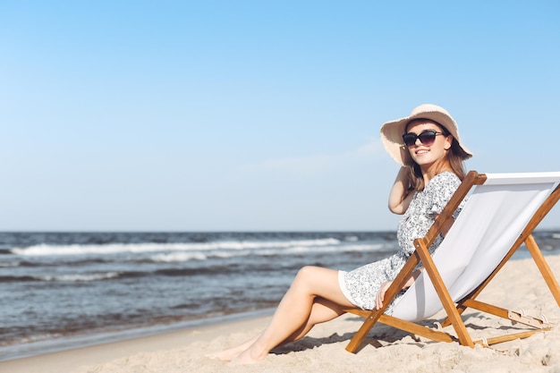 Donna castana felice che indossa occhiali da sole e cappello che si rilassa su una sedia a sdraio in legno sulla spiaggia dell'oceano.