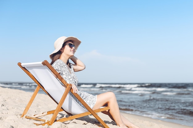 Donna castana felice che indossa occhiali da sole e cappello che si rilassa su una sedia a sdraio in legno sulla spiaggia dell'oceano.