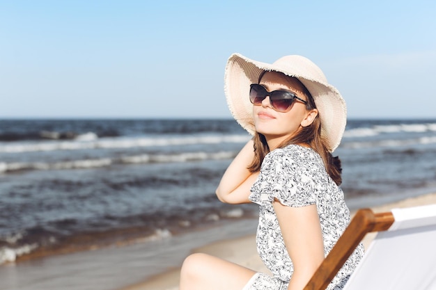 Donna castana felice che indossa occhiali da sole e cappello che si rilassa su una sedia a sdraio in legno sulla spiaggia dell'oceano.