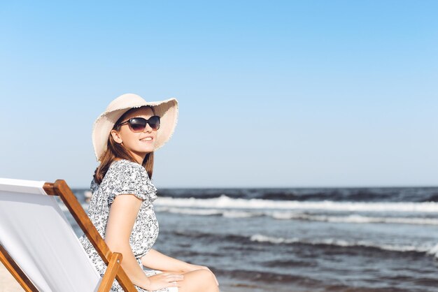 Donna castana felice che indossa occhiali da sole mentre si rilassa su una sedia a sdraio in legno sulla spiaggia dell'oceano.