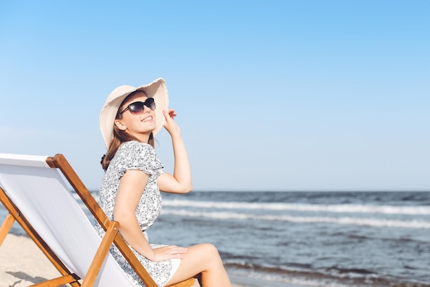 Donna castana felice che indossa occhiali da sole mentre si rilassa su una sedia a sdraio in legno sulla spiaggia dell'oceano.