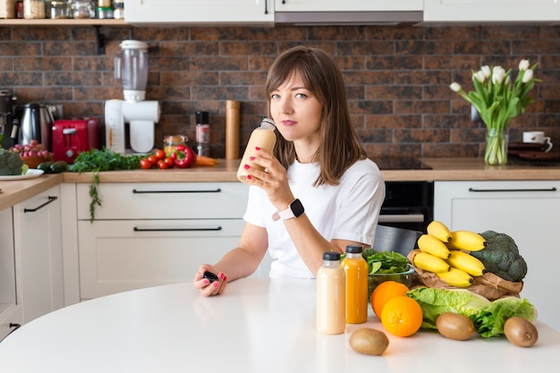 Donna castana felice che si siede con una bottiglia di frullato e frutta a casa cucina. Pasto vegano e concetto di disintossicazione. Ragazza con la maglietta bianca che beve cocktail fresco. Mockup di imballaggio