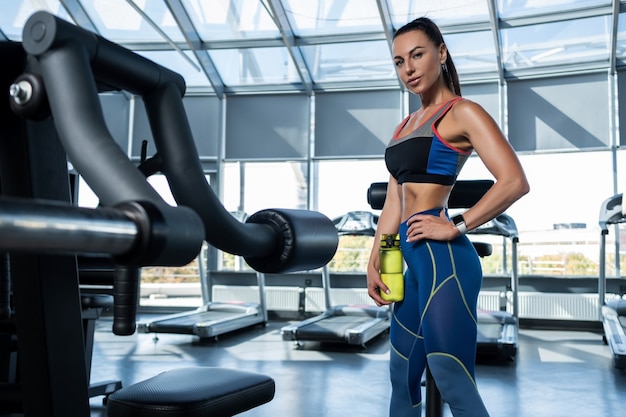 Donna castana in forma in piedi in palestra con una bottiglia d'acqua