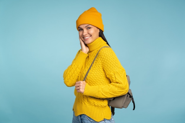 Donna castana in maglione lavorato a maglia giallo e cappello che sorride mentre sta in piedi con il suo zaino. Persona di sesso femminile in vestiti caldi in piedi sullo sfondo blu. Concetto invernale