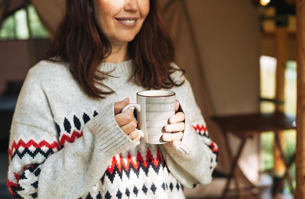 Donna castana in maglione nordico che beve tè e si rilassa in glamping nella natura