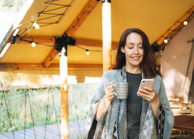 Donna castana in poncho che utilizza il telefono cellulare e si rilassa nel glamping nella natura