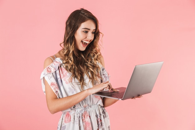 donna castana in vestito che tiene e che utilizza computer portatile d'argento, isolato sul colore rosa