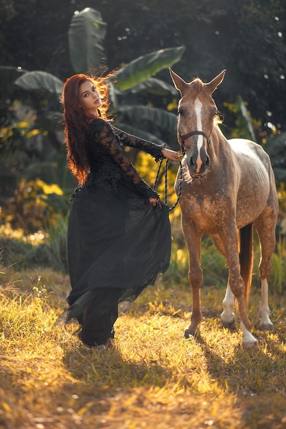 Donna castana in vestito nero con il cavallo nella foresta soleggiata di estate