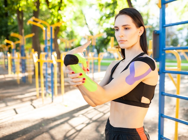 Donna castana muscolare sbalorditiva flessibile che indossa l'attrezzatura sportiva nera, allungando il braccio. Giovane atleta femminile fiducioso che pratica ginnastica, riscaldamento, kinesiotaping colorato sul corpo.