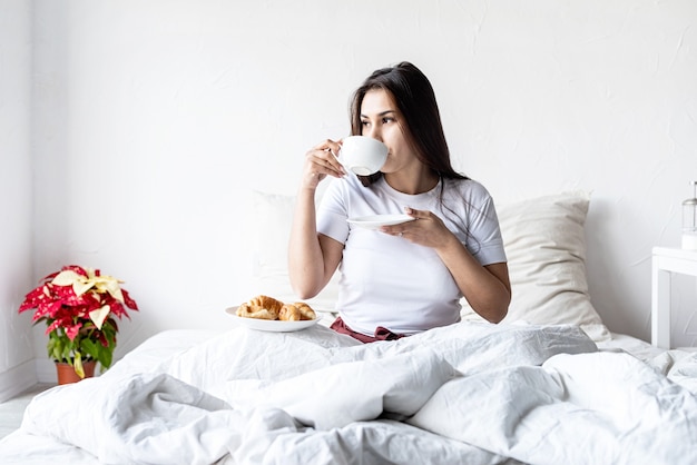 donna castana seduta sveglia nel letto con palloncini a forma di cuore rosso e decorazioni che bevono caffè mangiando croissant