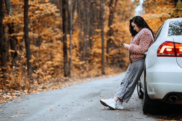 Donna castana sicura in piedi vicino all'auto e chiama il telefono Preparare la tua auto per l'autunno stagione autunnale Suggerimenti per la guida per l'autunno