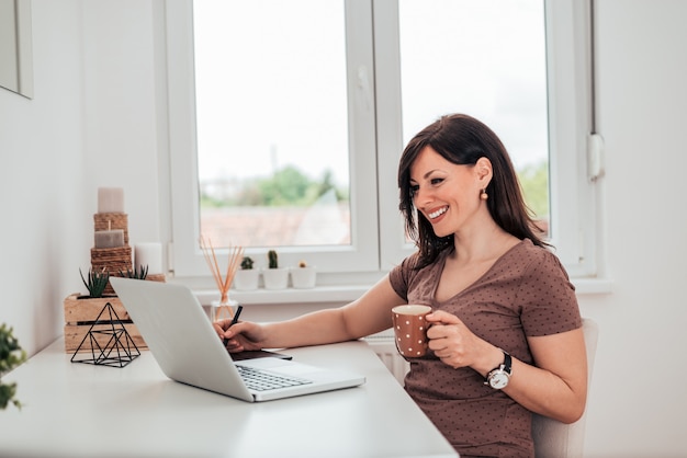 Donna casuale che beve il caffè di mattina e che utilizza computer portatile nell&#39;appartamento luminoso.