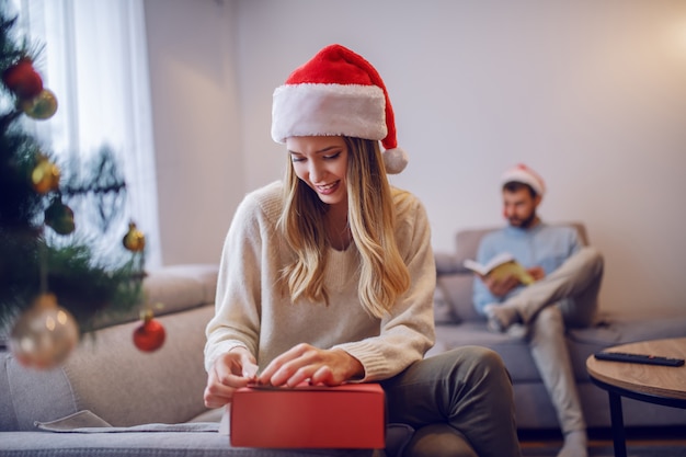 Donna caucasica attraente sorridente in maglione e con il cappello di Santa sulla testa che si siede sul sofà in salone accanto all'albero di Natale e ai regali dell'imballaggio. Sullo sfondo il suo ragazzo seduto e il libro di lettura.