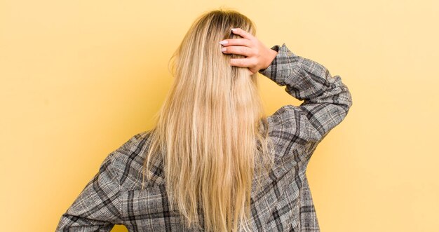 Donna caucasica bionda che si sente all'oscuro e confusa pensando a una soluzione con la mano sull'anca e l'altra sulla vista posteriore della testa