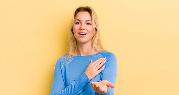 Donna caucasica bionda che si sente felice e innamorata che sorride con una mano accanto al cuore e l'altra allungata davanti