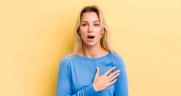 Donna caucasica bionda che si sente scioccata e sorpresa sorridendo prendendosi mano al cuore felice di essere quella giusta o di mostrare gratitudine