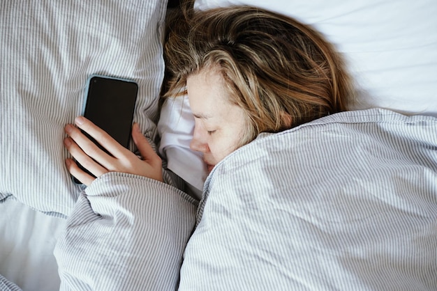 Donna caucasica che dorme a letto e tiene in mano lo smartphone Dipendenza online e concetto di social media