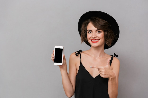 donna caucasica che indossa un abito nero e cappello che tiene smartphone, isolato sopra il muro grigio