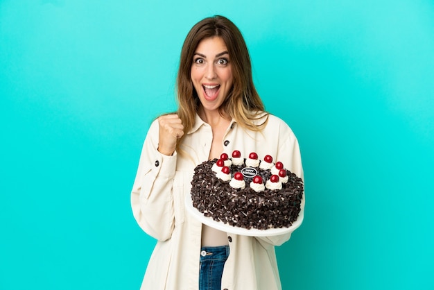 Donna caucasica che tiene la torta di compleanno isolata su fondo blu che celebra una vittoria nella posizione del vincitore